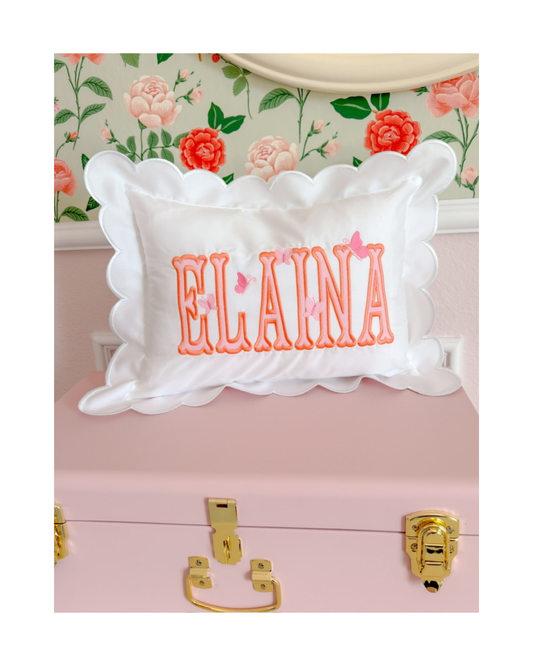 White pillow with 'ELAINA' embroidered on it, placed on a pink surface with floral wallpaper in the background.