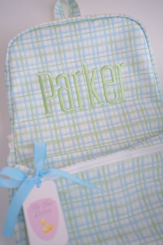 A close-up of a personalized TRVL backpack featuring a light blue and green plaid pattern, with the name 'Parker' embroidered in light green on the front. A light blue ribbon is tied to a white zipper, and a pink and white tag is attached.