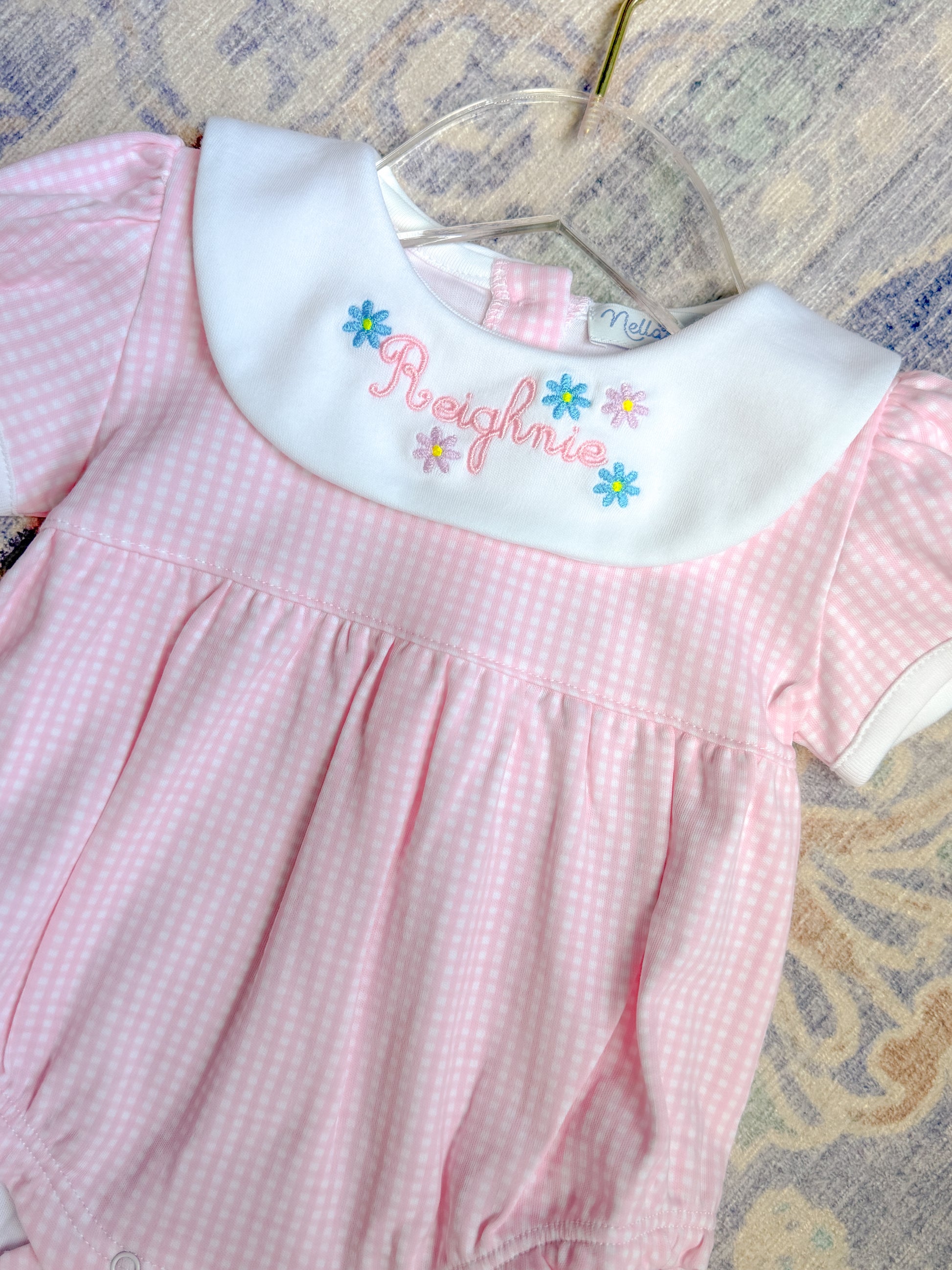 Pink checkered dress with embroidered name on a hanger against a patterned background