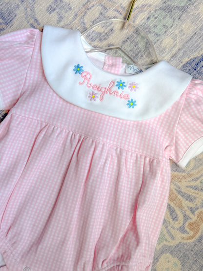 Pink checkered dress with embroidered name on a hanger against a patterned background